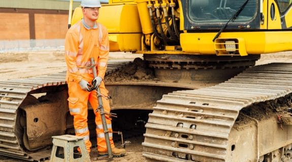 MATtest Site Services engineer performing bituminous testing beside heavy plant machinery on a construction site.